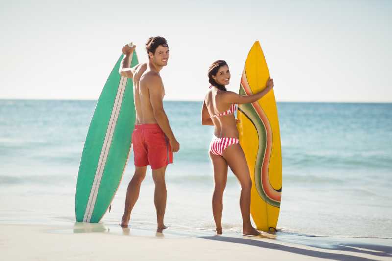 海滩上拿着不同颜色冲浪板回头笑的情侣