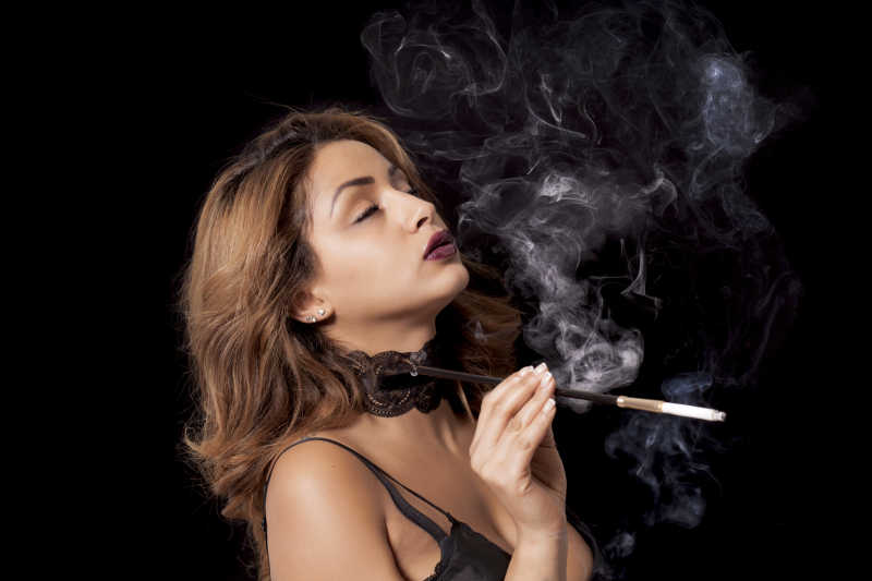 黑色背景下穿着性感内衣的拿着带烟嘴的香烟吸烟的美女