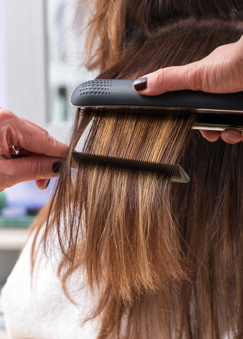 美发师在女客户的棕色长发上使用直发器