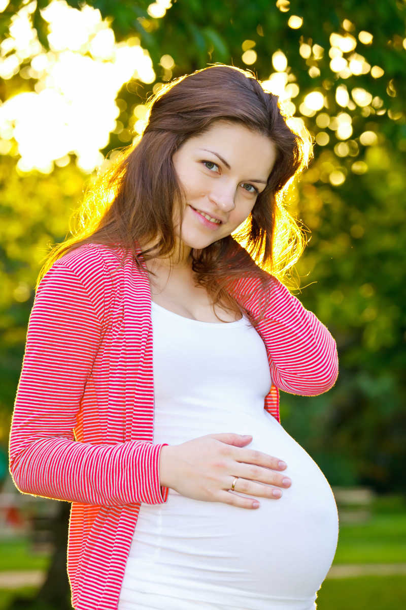 绿色有阳光的户外背景下摸着肚子的年轻孕妇