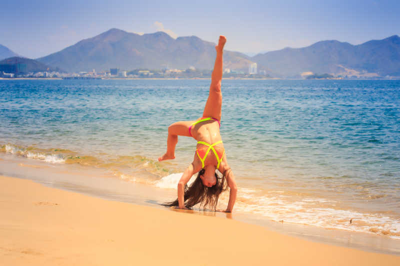 山峦背景下蓝色海边金色沙滩上穿着比基尼做瑜伽动作的美女