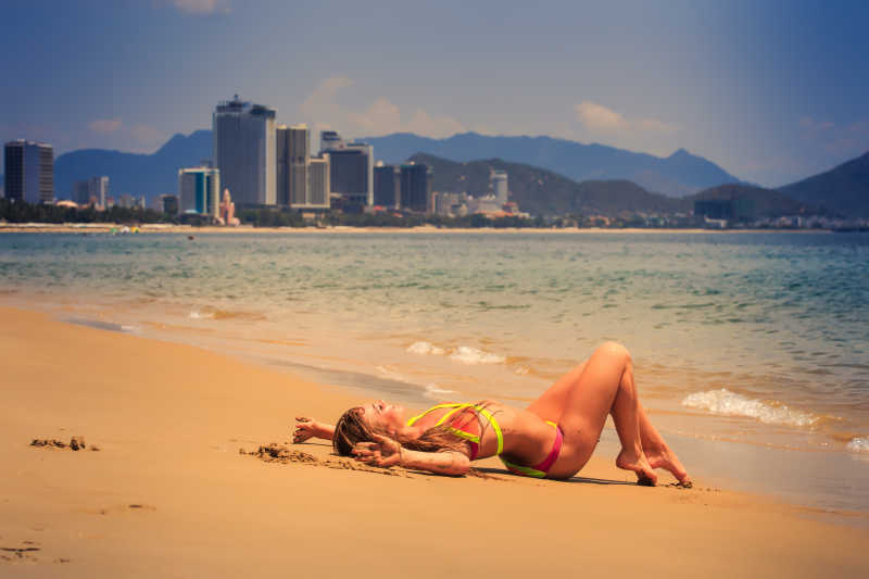 城市背景下海边穿着比基尼躺在黄色的沙滩上的美女