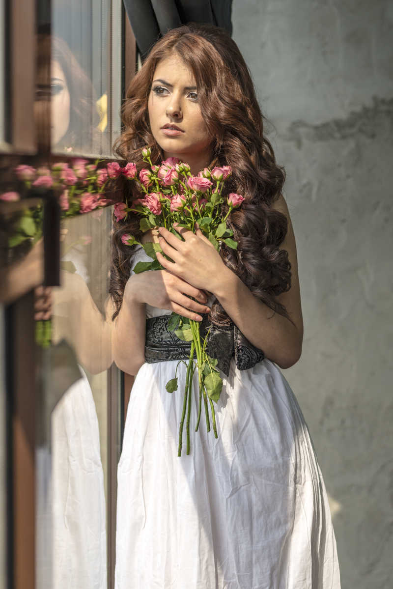 棕色卷发美女靠着窗拿着花