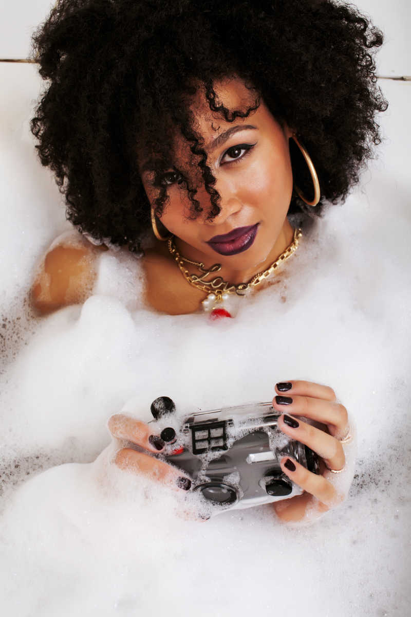 黑人少女躺在浴缸里泡着泡沫戴着无瑕疵的首饰拿着相机
