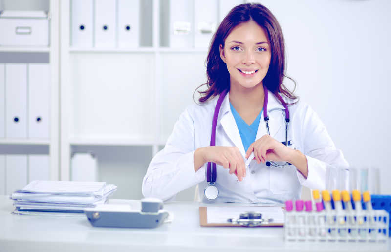 白色背景下坐在办公桌前拿着笔的微笑的女医生