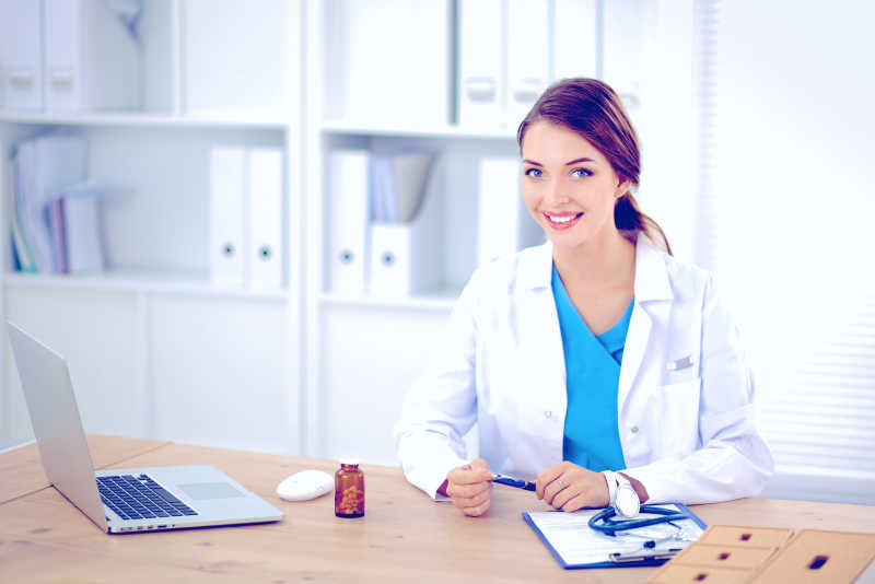 白色背景下拿着笔微笑着坐在办公桌前的女医生