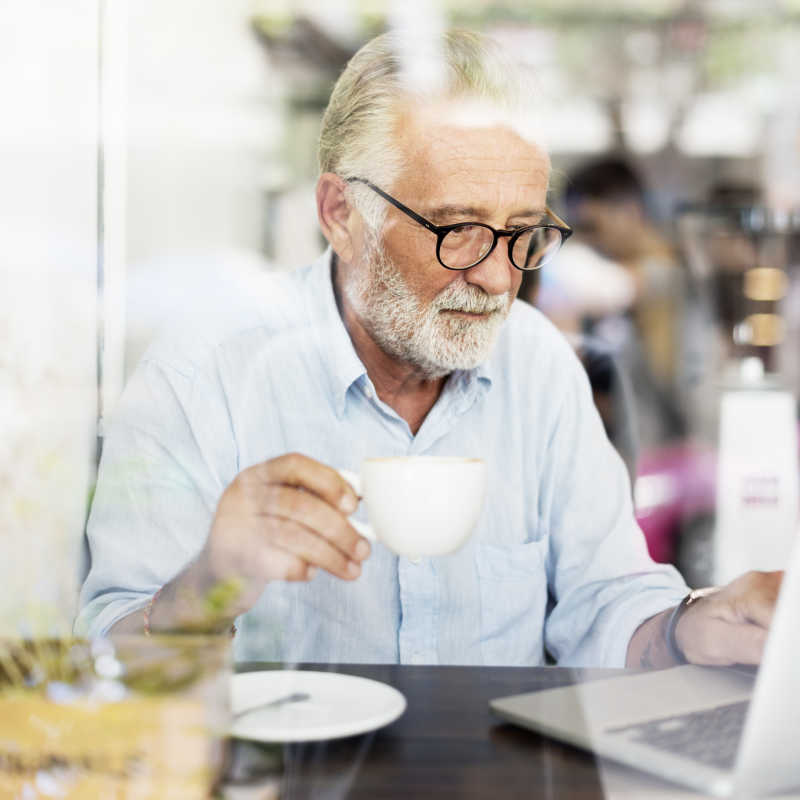 坐在咖啡馆使用笔记本电脑喝着咖啡的白胡子老商人
