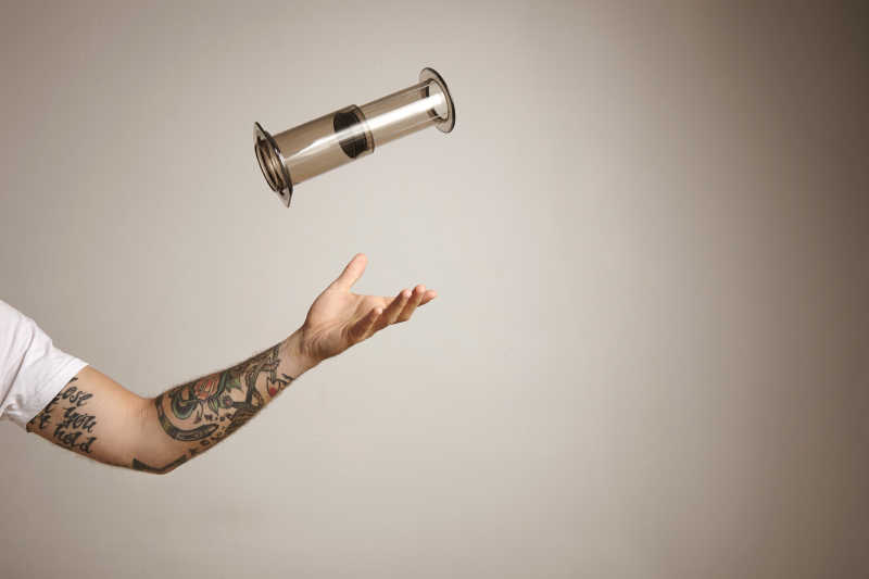 浅灰色背景下纹身的男人手臂在空中抛出一个小型咖啡机
