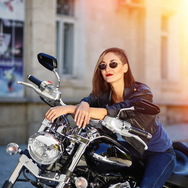 阳光下建筑旁边坐在摩托车上的穿黑色皮夹克的女孩