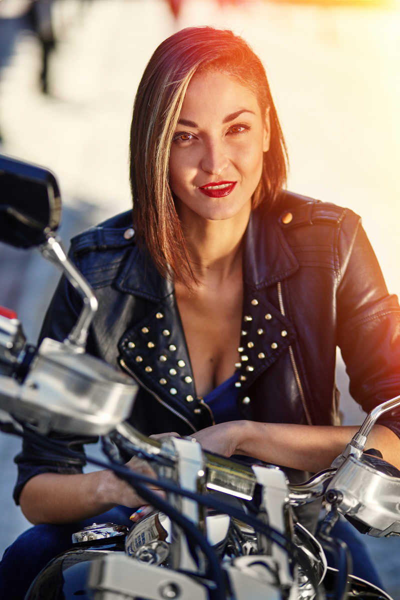 阳光下街道边坐在摩托车上的穿着黑色皮夹克的性感女孩