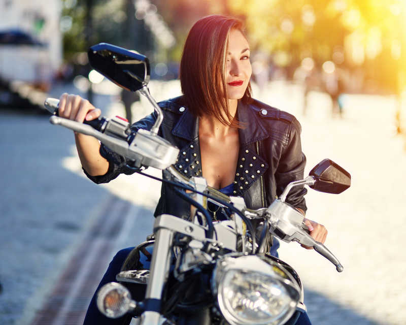 阳光下街道边坐在摩托车上的穿着黑色皮夹克微笑的女孩