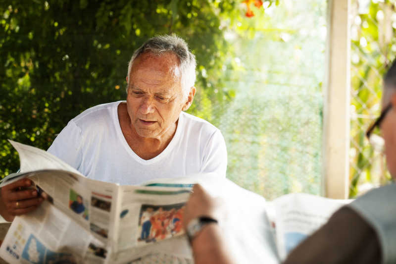 阳光下绿色植物背景下坐在一起看报纸的两位老年人