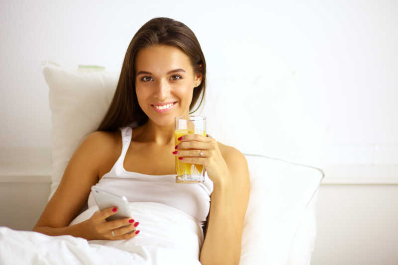 女人坐在床上喝果汁和玩手机
