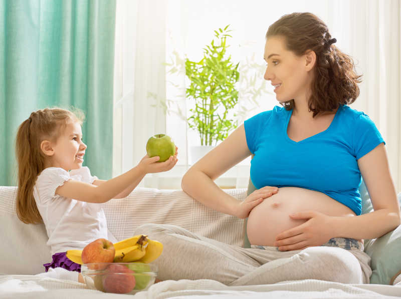 沙发上怀孕的妈妈和拿着水果的女儿