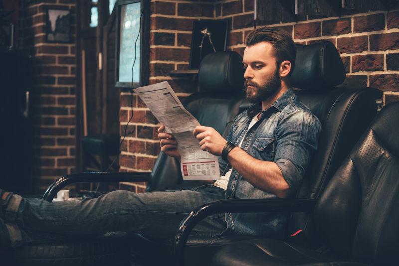 一个英俊的年轻胡子男子坐在理发店舒适的椅子上看报纸