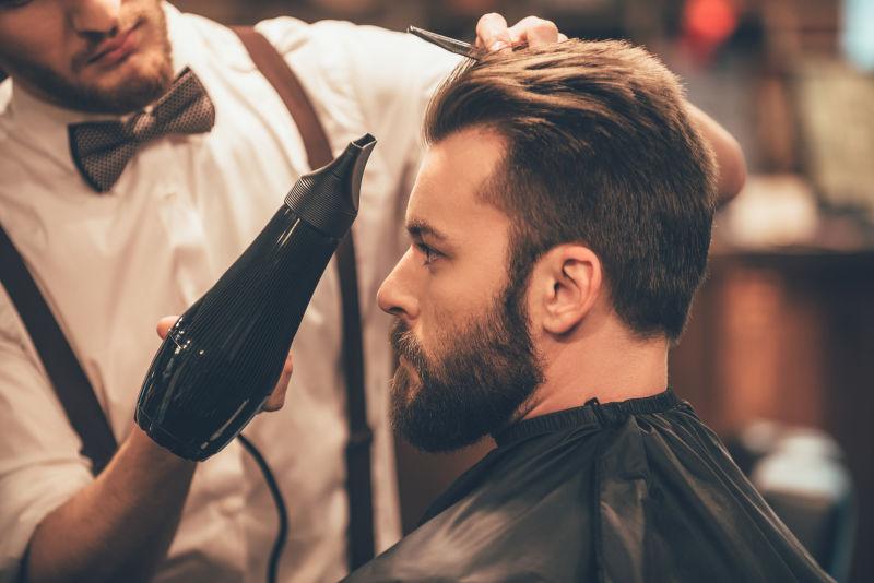 理发店理发师给年轻大胡子的男人用吹风机吹头发
