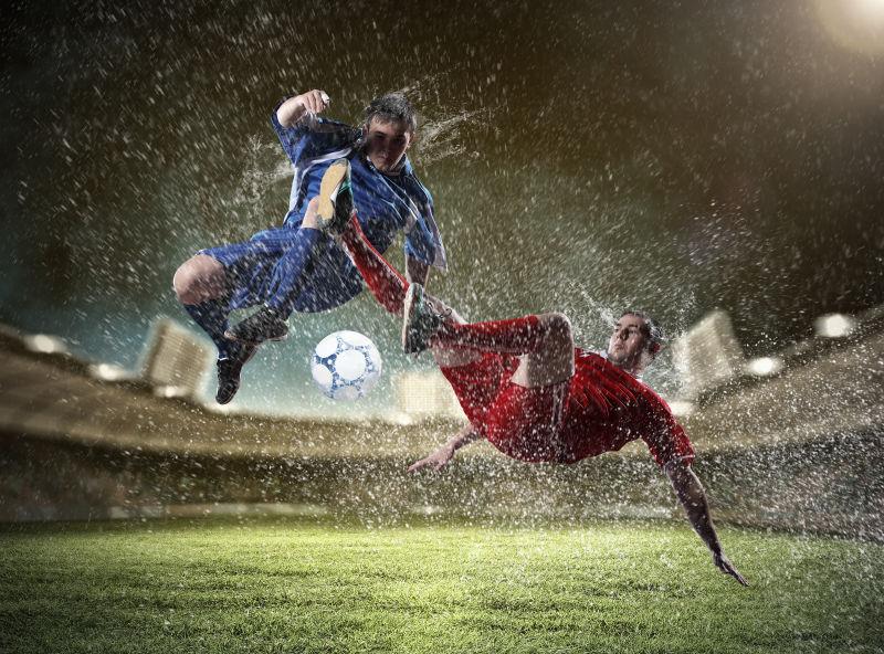夜晚的足球场上穿着红色和蓝色球服的两个足球运动员在抢球