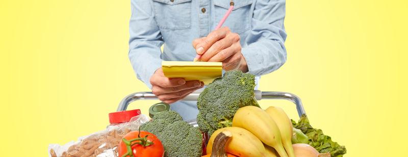 黄色背景下超市的员工记录购物车内的蔬菜和水果