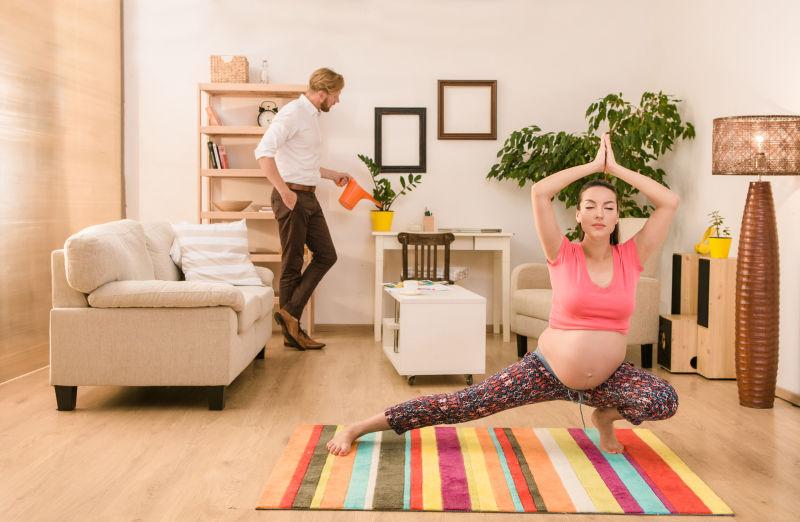 在家中练习瑜伽的孕妇和身后浇花的丈夫