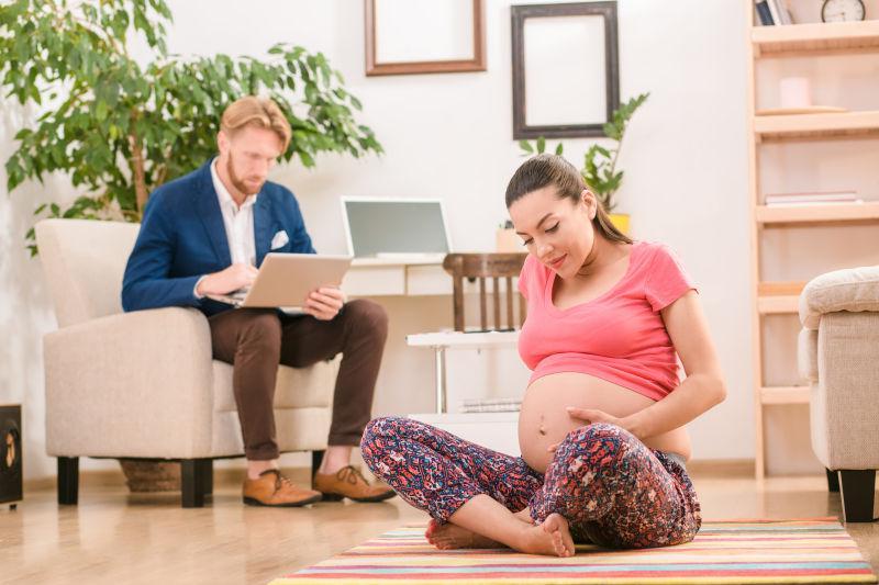 孕妇在做瑜伽是触摸腹部和她身后工作的丈夫