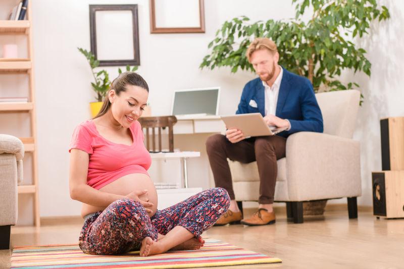 快乐的孕妈坐在瑜伽垫上抚摸着孕肚和沙发上工作的丈夫