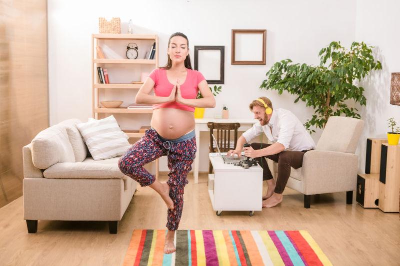 在家中练瑜伽的孕妇和坐在沙发上疯狂玩游戏的丈夫