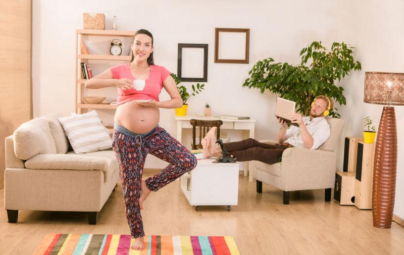 端着咖啡杯锻炼瑜伽姿势的孕妇和带着耳机看电脑的丈夫