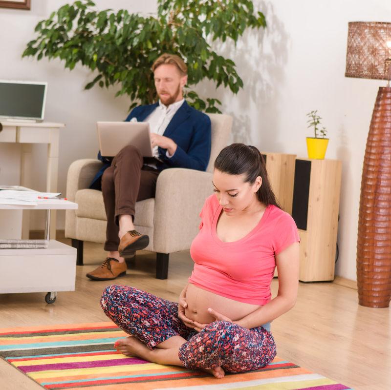 坐在瑜伽垫上的孕妇和沙发上坐着工作的丈夫