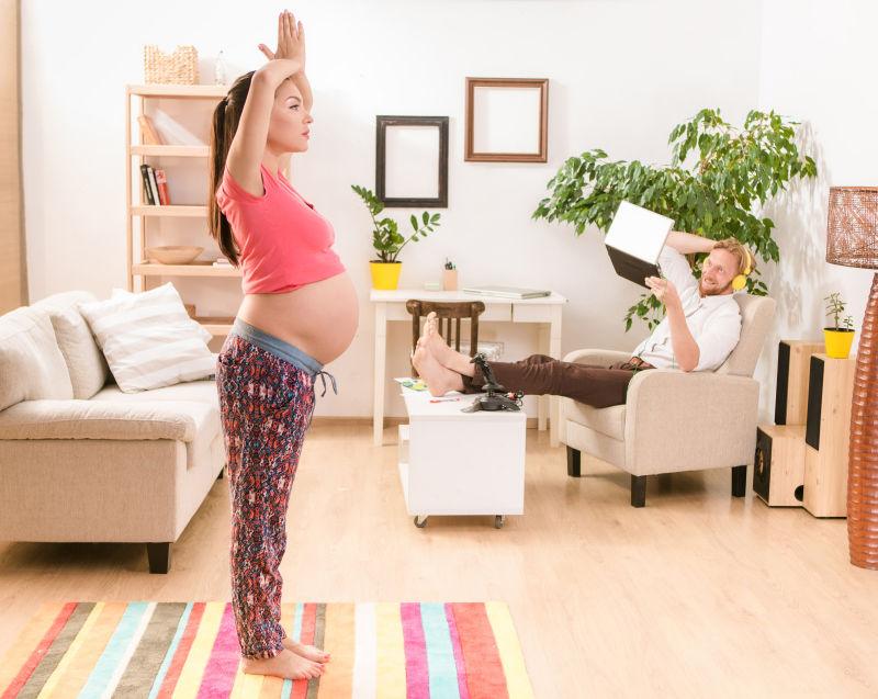在家中做瑜伽的孕妇和在沙发上拿着笔记本电脑开视频的丈夫
