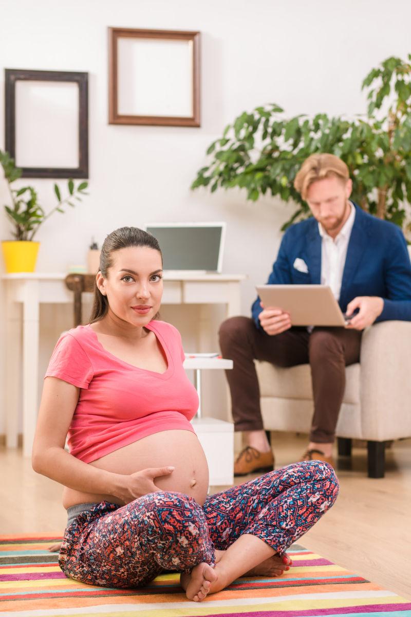 孕妇坐在瑜伽垫上和她身后正在工作的丈夫