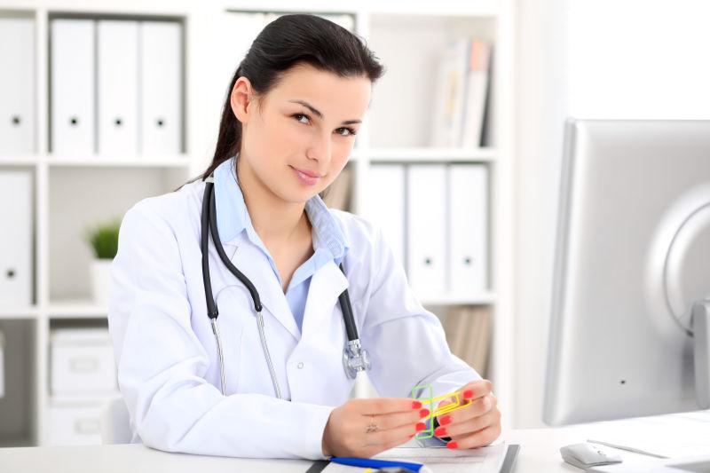 年轻的黑发女医生坐在桌子旁在医院的办公室里用电脑工作