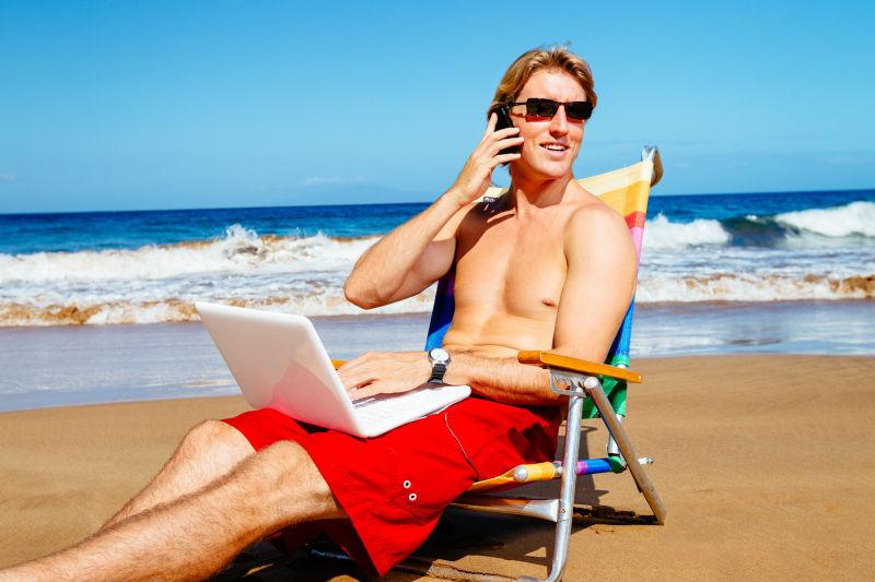 坐在海边沙滩椅上使用笔记本电脑和手机的年轻男人