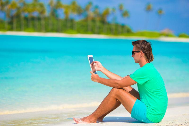 坐在热带沙滩上使用平板电脑的年轻男人