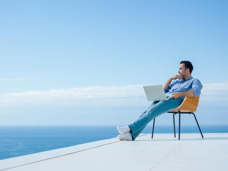 蓝天下坐在阳台上的拿着笔记本电脑的年轻男人