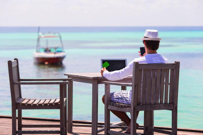 坐在海边木椅上使用手机和笔记本电脑的男人