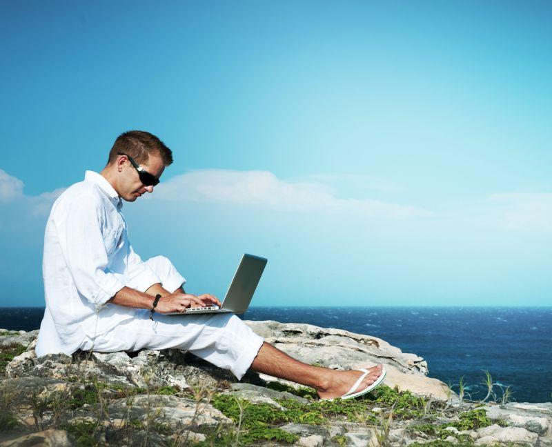 蓝天下坐在岩石上使用笔记本电脑的男人