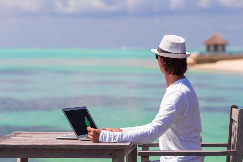 坐在海边木桌旁使用笔记本电脑的年轻人