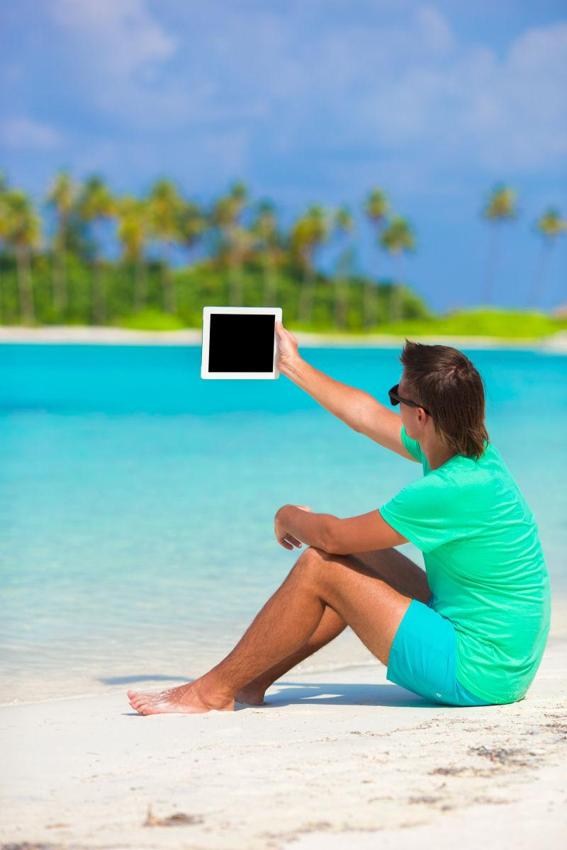 拿着平板电脑坐在海边沙滩上的男人