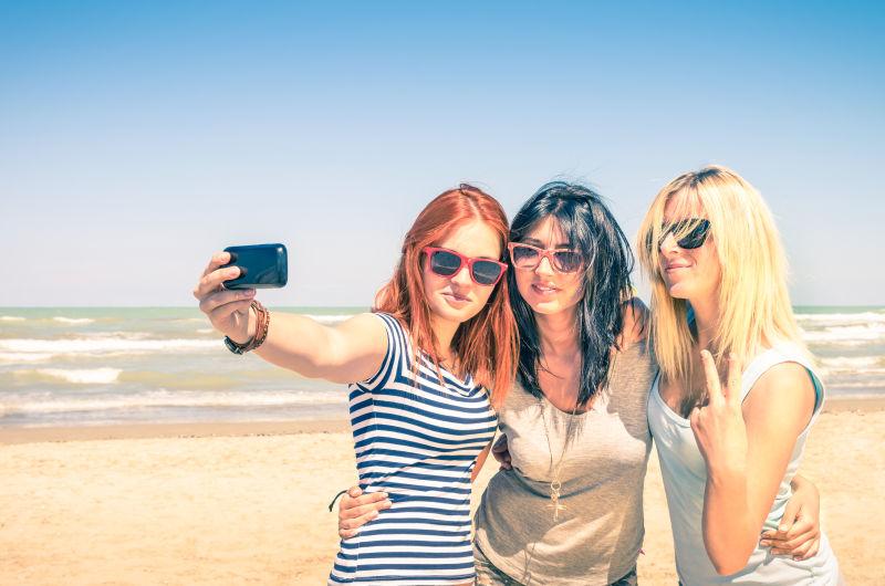 在海滩上自拍的女友们与现代智能手机一起享受美好时光