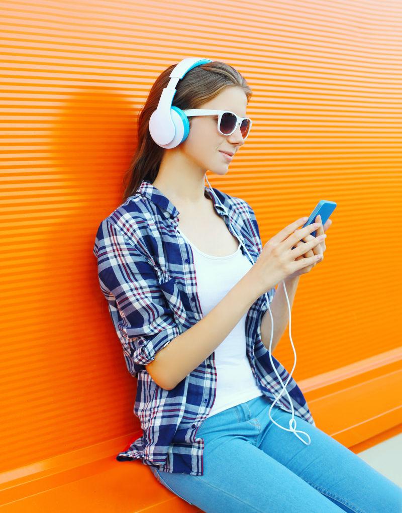 坐在橘色椅子上戴着耳麦使用智能手机听音乐的年轻女孩