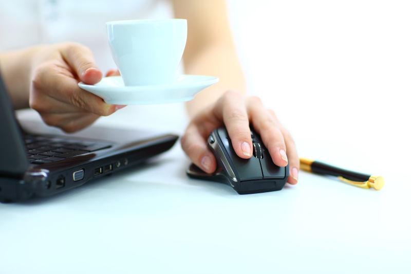 白色办公桌上一只手操控鼠标一只手端着咖啡的女人