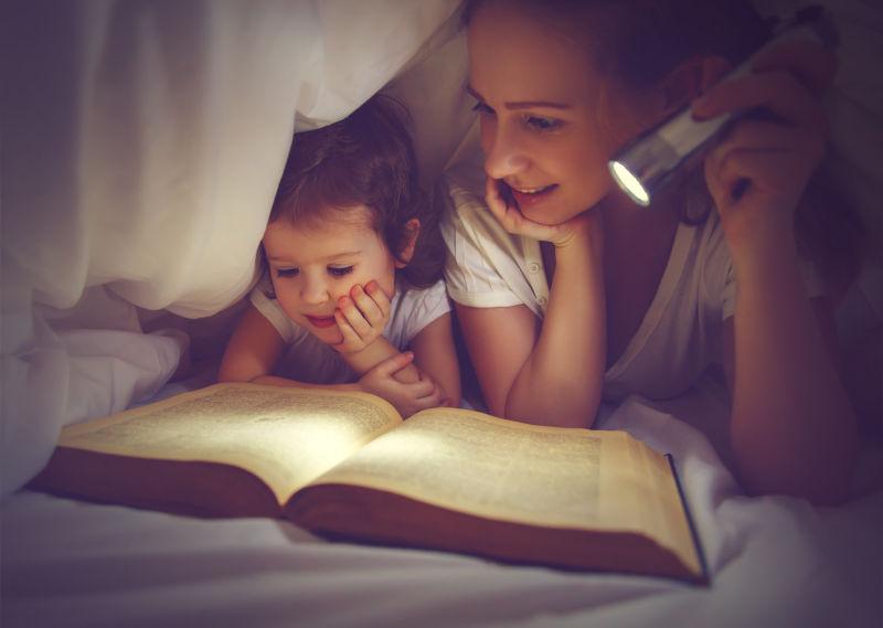 在床上妈妈和女儿用手电筒在毯子下看书