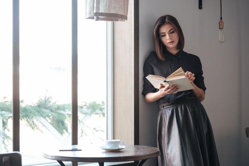 年轻美丽的女士站在咖啡馆窗户旁看书