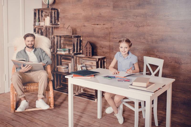 坐在书桌前写作业的小女孩和坐在摇椅里看书的父亲