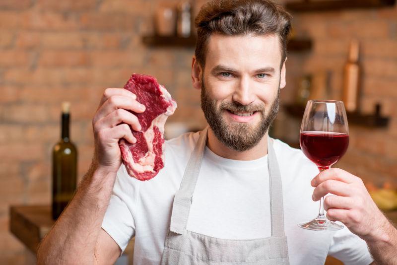 一个穿着围裙的英俊男子在厨房里拿着生肉和酒的肖像