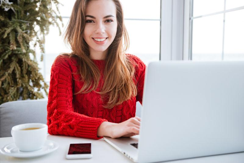 穿红毛衣的漂亮女士使用笔记本电脑身边放着手机和一杯茶
