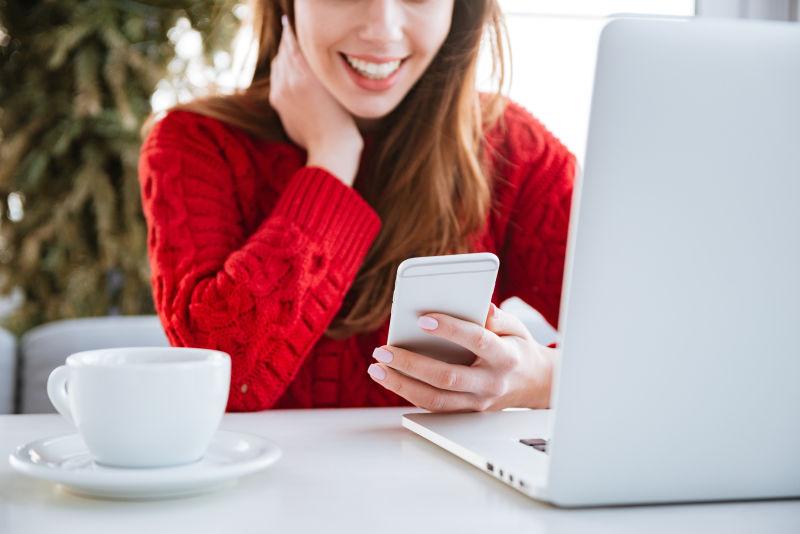 穿红毛衣的年轻美女使用手机聊天身前放着笔记本电脑和一杯茶