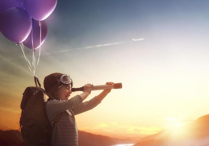 带旅行背包和气球的年轻孩子用望远镜看日落