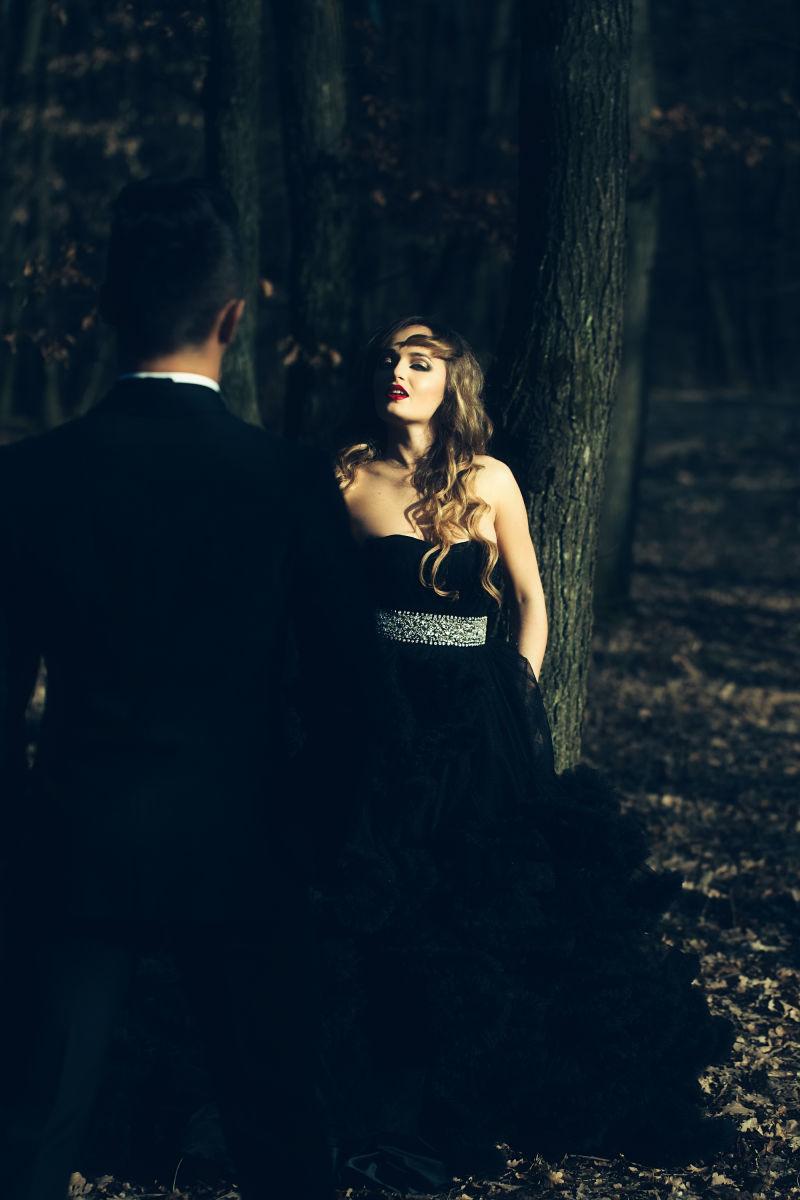 黑暗环境的森林中的英俊男子和身着一件漂亮晚礼服的性感女郎