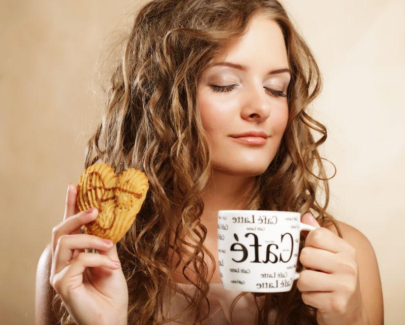 亚麻色背景下享受热咖啡和心型饼干的卷发美女